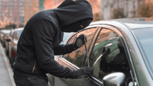 Pessoa com capuz preto assaltando um carro, representando um assalto a carro, para introduzir o assunto sobre proteção veicular furto e roubo.